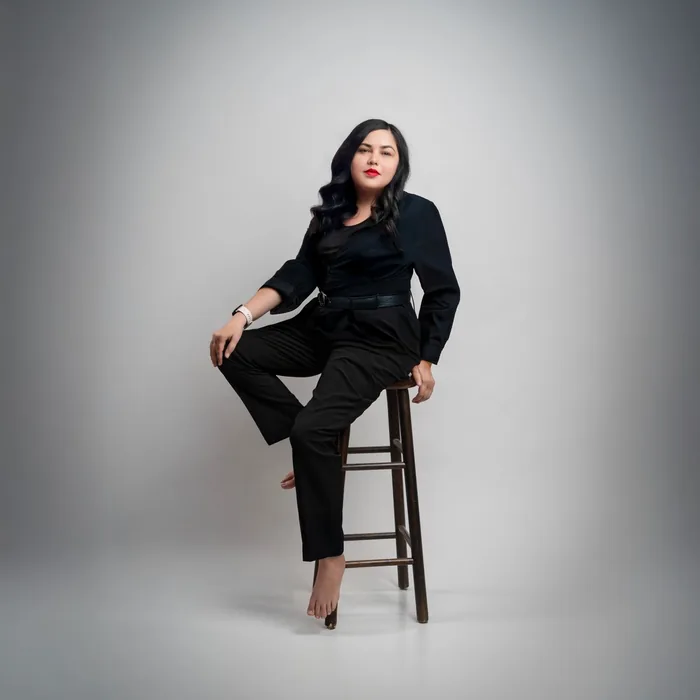 Elegant woman in black attire sitting on a stool against a gray studio background.